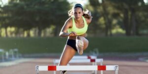 Female athlete in a neon green top clears a hurdle on an outdoor track during a race.